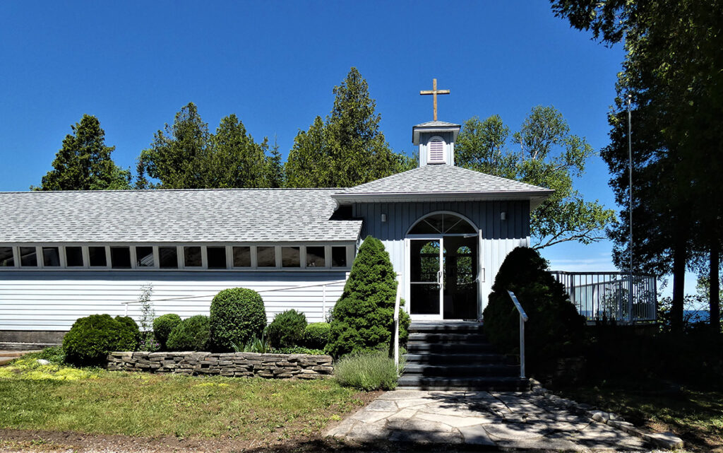Front view of St. Edmund’s Anglican Church in Tobermory, Ontario, featuring light blue wooden siding, a small bell tower with a cross, and surrounding greenery.